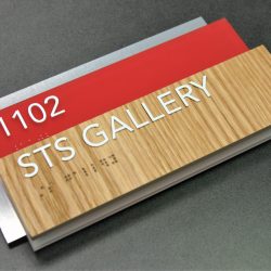 Backplate is .125” black acrylic with Chemetal laminate face. Mid-plate is .125” non-glare clear acrylic with subsurface paint with surface applied raised text and friction fit clear raster braille. Faceplate is .125” clear acrylic with WilsonArt laminate face with surface applied raised text and friction fit clear raster braille. STS Gallery